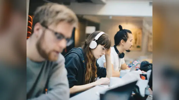 A woman with headphones rests her head on her hand, focused on a laptop, beside coworkers at a desk