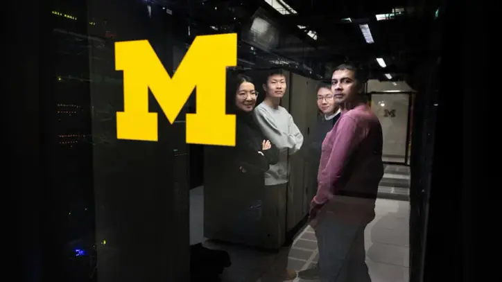 Three men and a woman stand in a circle between two rows of tall, black computer towers. They are looking through a glass panel stamped with the University of Michigan block M.