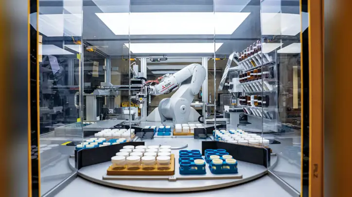 A large, white robotic arm works inside a glass enclosure, surrounded by trays of capped sample jars.